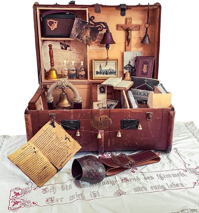 Ein alter Lederkoffer mit verschiedenen Gegenständen zum Thema Dom. Zum Beispiel ein Kruzifix, Glocken, Fotografien und Bücher.