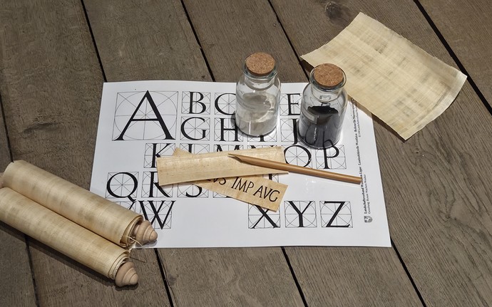 Auf einem Tisch liegen ein Bambusgriffel, Papyrusblätter und eine Papyrusrolle. Auf einem Blatt mit dem Alphabet stehen zwei Fläschchen mit Tinte.